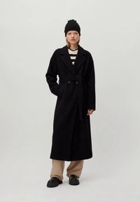 Donna che indossa un lungo cappotto nero con cintura, un berretto nero, pantaloni beige, scarpe nere e una maglietta a righe bianche e nere, in piedi di fronte a uno sfondo semplice.