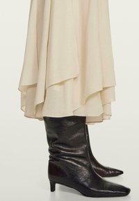 Zwarte leren kniehoge laarzen met een spitse neus en een lage hak, gecombineerd met een vloeiende beige rok met gelaagde ruches aan de onderrand.