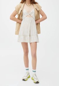Vestido a rayas blancas con un dobladillo con volantes, detalles de corte y lazo en el pecho, combinado con una chaqueta beige y zapatillas grises.