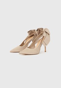 Μπεζ ψηλοτάκουνα με μύτη σε μυτερό σχήμα, με λουριά στον αστράγαλο και μεγάλα υφασμάτινα φιόγκια στο πίσω μέρος, πάνω σε λευκό φόντο.