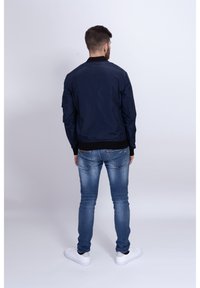 Námořnická bomber bunda s žebrovaným límcem, elastickými manžetami a pasem. V kombinaci s těsně padnoucími modrými džíny a bílými teniskami, pohled zezadu.