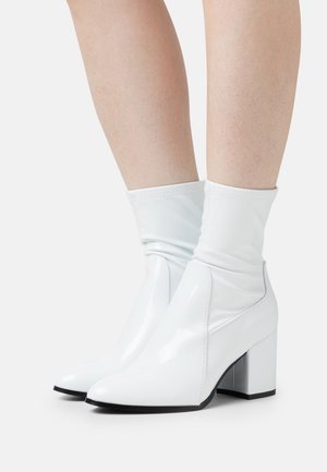Stivaletti alla caviglia in pelle verniciata bianca con una texture liscia, punta affusolata e un alto tacco a blocco. Il design presenta un risvolto morbido nella parte superiore.