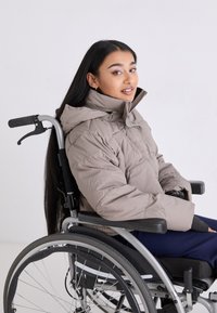 Padded beige jacket with high collar, featuring a zip closure and insulated design, worn by a person in a wheelchair.