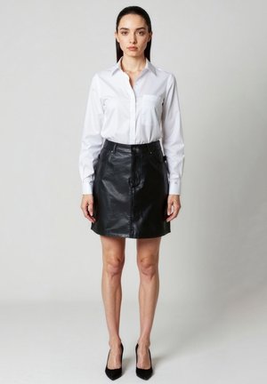 Frau steht frontal, trägt ein weißes Hemd mit Knöpfen, einen schwarzen Lederrock und schwarze hohe Absätze vor einem schlichten grauen Hintergrund.