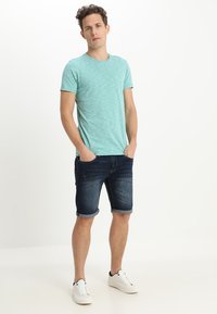 Camiseta de manga corta azul claro con un patrón texturizado, combinada con pantalones cortos de mezclilla oscura y zapatillas blancas. El modelo está de pie con las manos en los bolsillos.