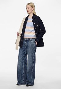 Blonde woman wearing a dark denim jacket, pastel striped sweater, wide-leg blue jeans, holding a wide beige belt across her shoulder.