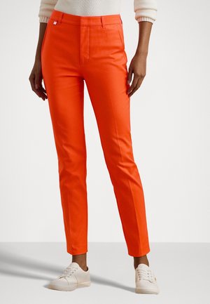 Personne portant un pantalon slim orange vif avec des poches avant et des baskets blanches, debout devant un fond blanc uni.