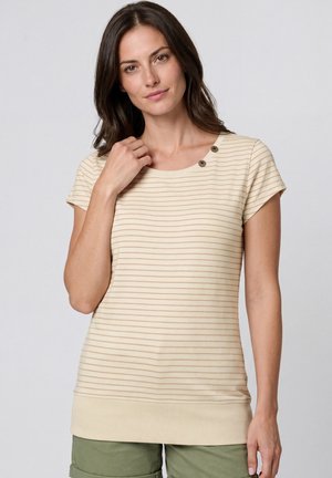 Frau mit dunklen Haaren, die ein beige gestreiftes Kurzarmoberteil mit Knopfdetails und olivgrüne Shorts trägt und vor einem einfarbigen Hintergrund posiert.