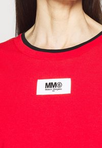 Röd bomullströja med rund svart krage och en rektangulär vit etikett som visar varumärket "MM6 Maison Margiela" i svart text.