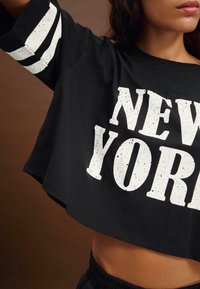 Černá zkrácená mikina s nápisem "NEW YORK" v bílém žíhaném tisku. Disponuje bílými pruhovanými detaily na rukávech. Měkká tkanina, pohodlný střih.