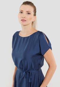 Laivansinisat satinimekko, jossa on lyhyet hihat, pyöreä pääntie ja vyötärön solmimisdetalji. Silkkinen pinta ja kevyt kiilto lisäävät sen eleganssia.