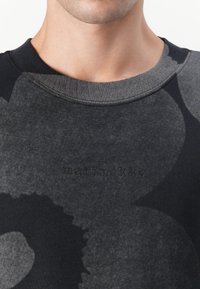 Schwarzer Pullover mit großen grauen Blumenmustern, strukturiertem Stoff und geripptem Kragen. Markenname "marimekko" in der Mitte eingeprägt.