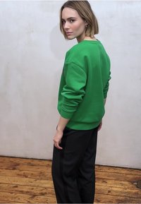 Vihreä collegepaita, jossa on rento istuvuus ja resorit hihoissa, yhdistetty mustiin housuihin, joissa on sivuraidat, puulattian taustalla.