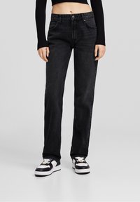 Black straight-leg jeans with a mid-rise waist, five pockets, and subtle stitching details, paired with black and white sneakers.