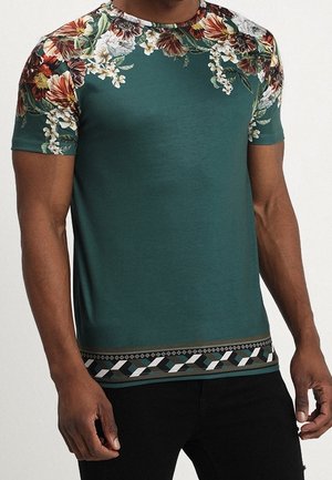 Mann trägt ein grünes kurzärmeliges Hemd mit Blumenmustern auf den Schultern und einem geometrischen Muster nahe dem Saum, kombiniert mit schwarzer Hose.