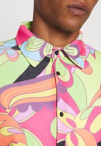 Camisa de manga corta con un patrón abstracto en espiral colorido. Presenta un cuello verde, botones negros y una tela ligera y texturizada.