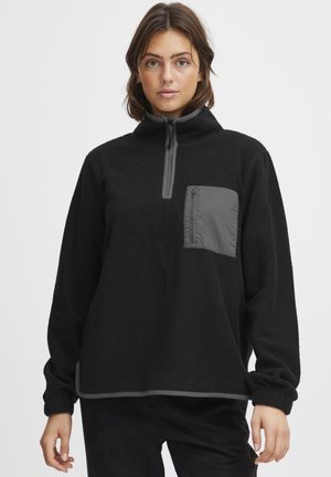 Svart fleece-pullover med høy krage, halv glidelås foran og en grå glidelåslomme på brystet. Har elastiske mansjetter og søm.