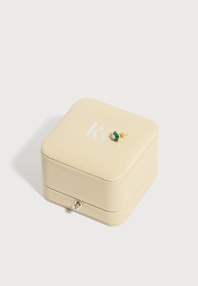 Μικρό σκουλαρίκι με χρυσό και πράσινο πολύτιμο λίθο, το οποίο ακουμπά σε κλειστό μπεζ κουτί κοσμημάτων με ασημένιο κούμπωμα και μερικά λευκά γράμματα.