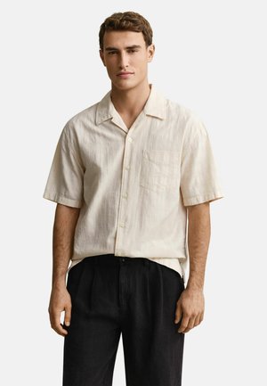Jeune homme portant une chemise crème à manches courtes avec poche et un pantalon noir, debout devant un fond clair uni.