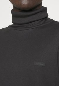 Maglione a collo alto nero con texture a costine e design aderente. Presenta un piccolo tag rettangolare con il logo Calvin Klein sul davanti.