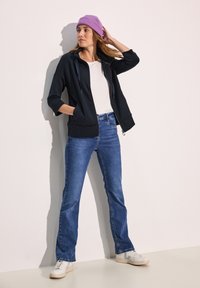 Felpa nera con zip e cappuccio regolabile, abbinata a jeans blu e sneakers bianche. Un berretto a coste color lavanda aggiunge un tocco di colore.