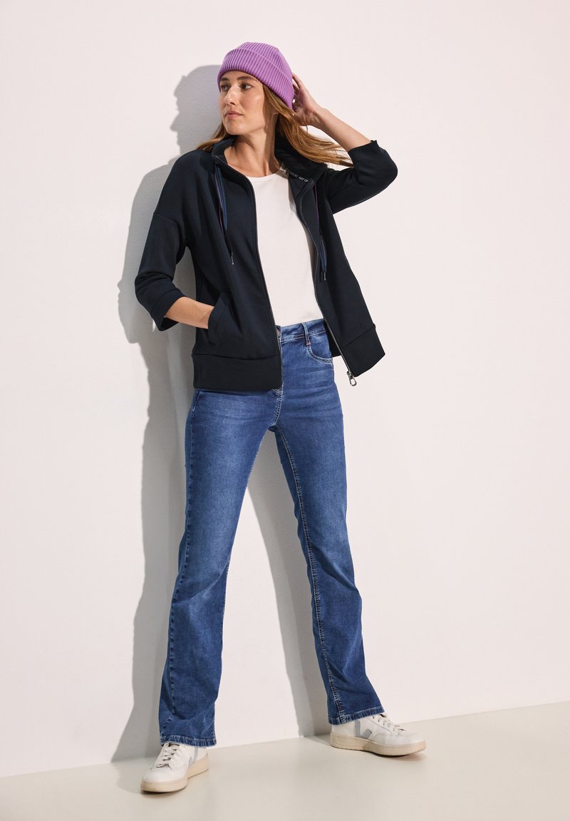Felpa nera con zip e cappuccio regolabile, abbinata a jeans blu e sneakers bianche. Un berretto a coste color lavanda aggiunge un tocco di colore.