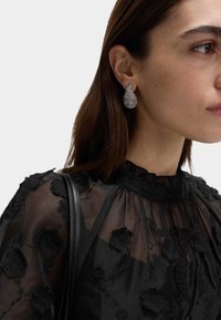 Kolczyki w kształcie kropli łzy z fakturowaną powierzchnią ze srebra, noszone z czarną bluzką w kwiatowy wzór.