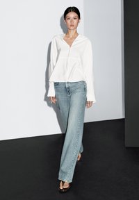 Hvid, langærmet, knappet bluse kombineret med lyseblå bukser med vide ben, suppleret med brune sandaler med slidser. Enkel, ren design.