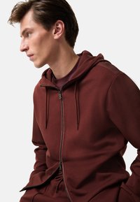 Bordeaux rode rits hoodie met een trekkoordkap, zachte textuur en eenvoudig ontwerp. Beschikt over een zichtbare rits en ribgebreide accenten bij de manchetten.