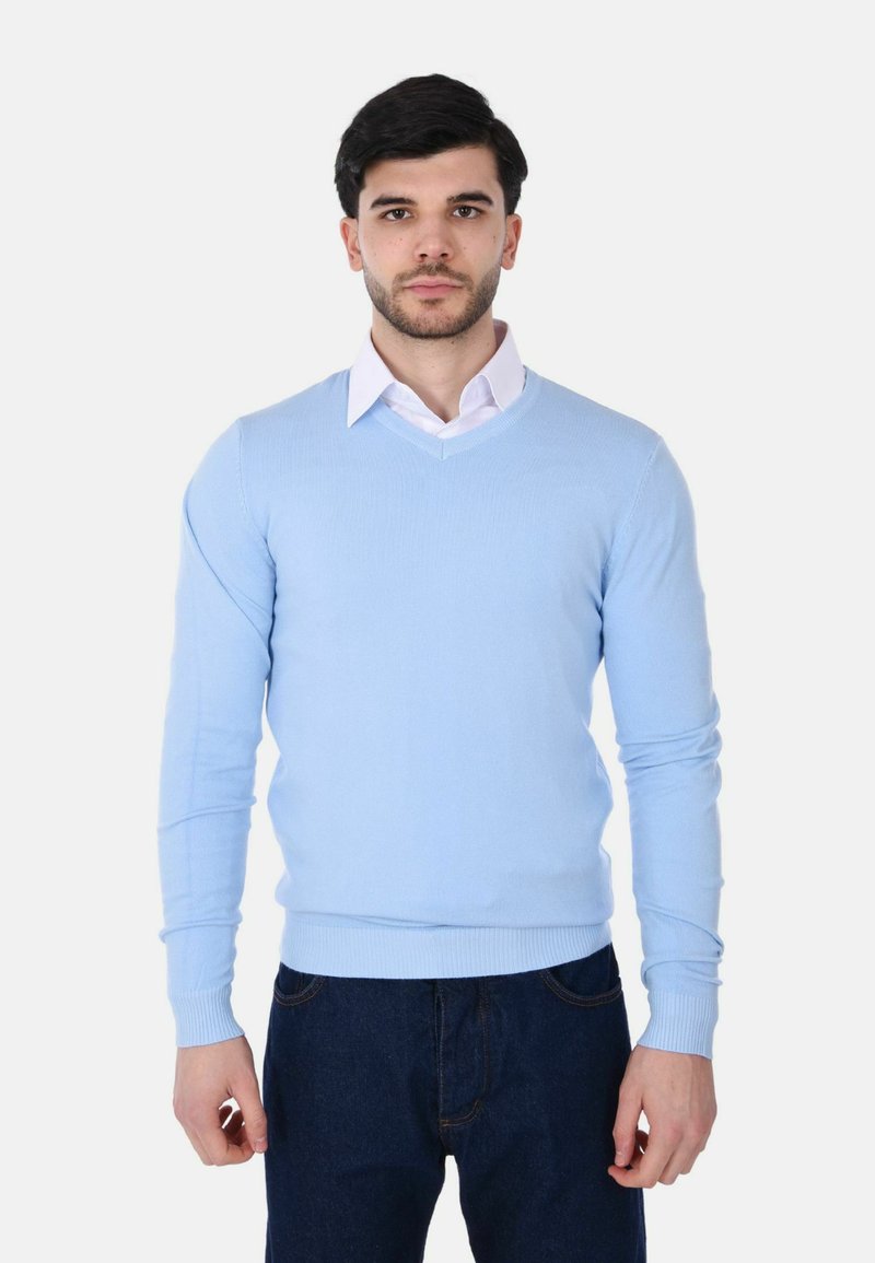 Uomo che indossa un maglione azzurro chiaro a V sopra una camicia bianca con colletto e jeans blu scuro, in piedi contro uno sfondo bianco semplice.