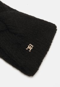 Fascia nera lavorata a maglia realizzata in materiale morbido, caratterizzata da un accento in metallo dorato con il logo. La trama appare pelosa e spessa.