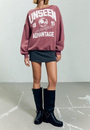 Osoba ubrana w oversize'ową bordową bluzę z nadrukiem „UNSEEN 1991 Michigan ADVANTAGE”, krótką czarną spódnicę i wysokie czarne kozaki.