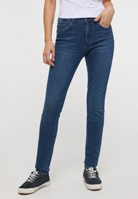 Tmavomodré skinny džínsy vyrobené z denimu, s piatimi vreckami, zapínaním na gombík a zips a hladkou textúrou. Nosia sa s čiernymi teniskami.