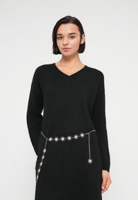Μαύρο πλεκτό φόρεμα με V-λαιμόκοψη, μακριά μανίκια και ασημένια αλυσίδα με μοτίβο αστέρια στη μέση. Μαλακή υφή και άνετη εφαρμογή.