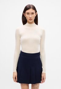 Blusa de gola alta em creme com textura canelada, combinada com uma saia plissada em azul-marinho com detalhes de botão lateral e um pequeno logótipo na cintura.