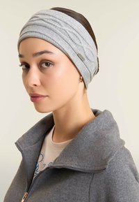 Fascia grigia in maglia con un motivo testurizzato. Indossata con una giacca grigia con zip, che mette in evidenza un materiale liscio e morbido sulla pelle.