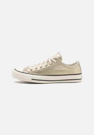 Zapatilla de lona dorada con punta de goma blanca, cordones y suela rayada en negro. Presenta un brillo metálico y un diseño clásico de corte bajo.