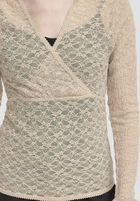 Beige Spitzenoberteil mit Wickeldesign, mit floralem Muster, langen Ärmeln und gewelltem Saum. Untere Lage in Schwarz sichtbar.