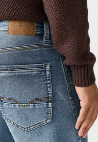 Džinsi no denima ar izbalinātu zilu krāsu un kontrastējošu oranžu šuvju. Iespējojams brūns ādas uzlīme ar uzrakstu "DENIM SUPPLY" un teksturēts brūns džemperis.