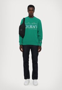 Grøn sweatshirt med teksten "ICECREAM" og skyline-grafik, parret med mørke jeans og hvide sneakers. Inkluderer også en sort crossbody-taske.