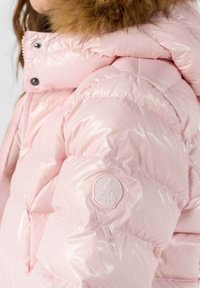 Chaqueta acolchada brillante en rosa con capucha con borde de piel, luciendo un parche de logo en la manga que presenta a un ciclista y el texto "JOTT".