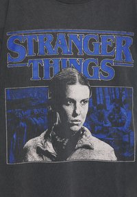 Schwarzes Baumwoll-T-Shirt mit dem Schriftzug "Stranger Things" in Blau, dazu eine Grafik mit dem Gesicht eines Charakters und einer Hintergrundszene in Blautönen.