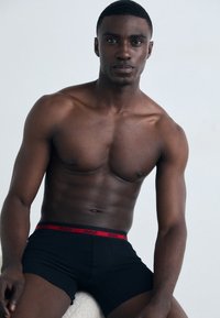 Schwarze Boxer Shorts mit einem roten "HUGO" Logo-Bund, die einen engen Sitz und elastisches Material bieten. Das Modell sitzt vor einem hellen Hintergrund.