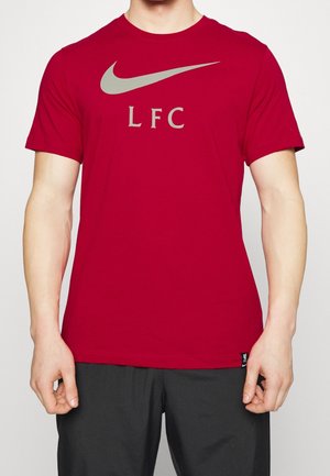 Homme portant un t-shirt rouge Nike avec un logo swoosh gris et le texte "LFC", associé à un pantalon noir, torse visible, fond neutre.