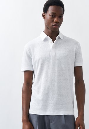 Άνδρας που στέκεται μπροστά σε απλό φόντο, φοράει λευκό πόλο μπλουζάκι με κοντά μανίκια και γκρι παντελόνι, κοιτάζοντας ελαφρώς προς τα κάτω.