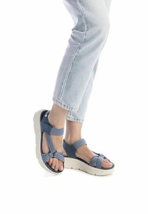 Sandalo blu con più cinturini, caratterizzato da una spessa suola bianca, tessuto testurizzato e design open toe. Indossato con jeans tagliati blu chiaro.