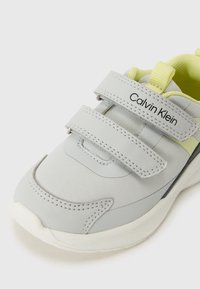 Light gray athletic shoe with yellow accents, featuring Velcro straps, a padded collar, rounded toe, and textured white sole.