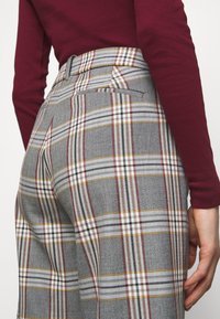 Grå rutiga byxor har hög midja med beige, röd och svart mönster. En bakficka syns nära midjebandet.