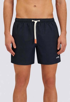 Uomo con pantaloncini da bagno neri con cordoncino bianco e punte arancioni, in piedi con le braccia rilassate lungo i fianchi.