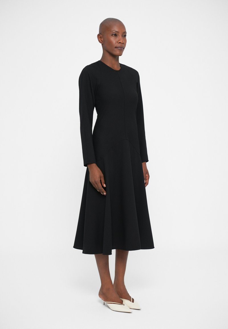 Femme portant une robe midi noire à manches longues avec une jupe évasée et des escarpins à bout pointu blancs, se tenant devant un fond uni.